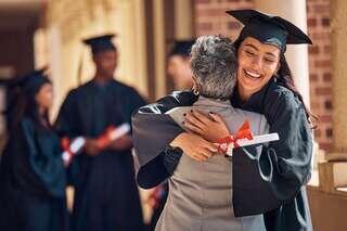 A smiling graduate in cap and gown hugs a family member while holding a diploma, celebrating a milestone achievement at a graduation ceremony.