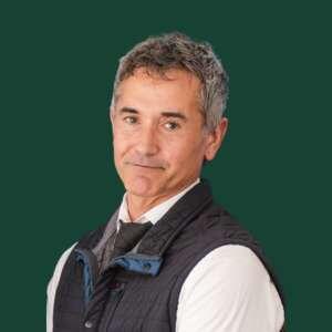 A middle-aged man with gray hair and a slight smile, dressed in a vest and tie, against a green background.