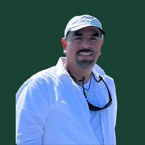 A man with a mustache smiles while wearing a white shirt and baseball cap, standing against a solid dark green background.