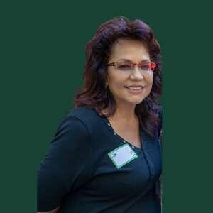 A woman with long, dark hair, wearing glasses and a black top, smiles with a nametag visible on her side against a green backdrop.