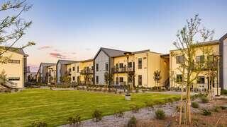 Modern residential buildings surround a manicured lawn, with trees and shrubs creating a welcoming, landscaped outdoor space at sunset.