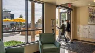 A modern lobby features a green chair, large windows, and sunlight streaming in, while two people walk through an entrance to an outdoor area.