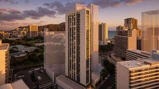 A modern high-rise amidst urban buildings, with a sunset backdrop illuminating the coastal landscape and distant mountains.