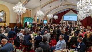 A formal gathering in an elegant banquet hall with attendees seated at tables, featuring chandeliers and a speaker addressing the audience.