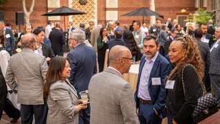 A networking event with professionals mingling outdoors, dressed in business attire, engaged in conversation amid tables and umbrellas.