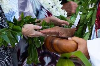 Hands exchanging a ceremonial bowl, adorned with greenery and floral leis, signify a moment of connection and tradition.