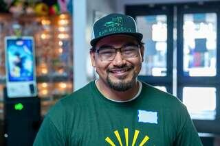 A smiling man with glasses and a cap stands in a bright setting, wearing a green shirt and a name tag. Colorful decorations are in the background.