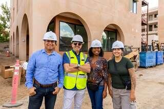A diverse group of four construction professionals, wearing hard hats and safety vests, pose together at a building site.