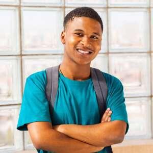 A smiling young man stands confidently with arms crossed, wearing a turquoise t-shirt and a backpack, against a bright window backdrop.