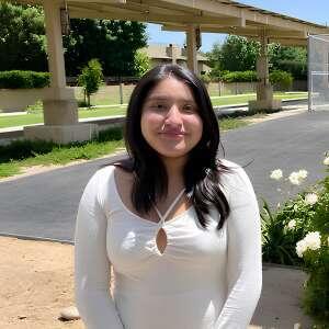 A young woman stands outdoors, smiling, wearing a white top, with green grass and trees in the background under sunny skies.