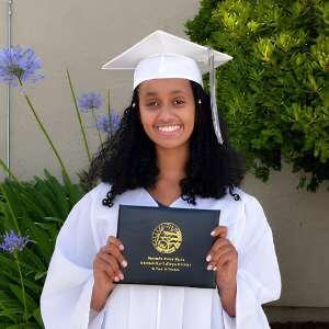 A proud graduate in a white cap and gown smiles while holding a diploma, surrounded by greenery and flowers.