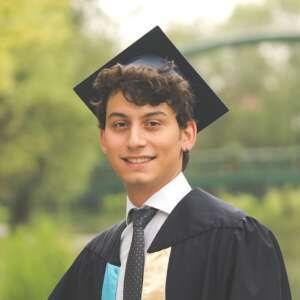 A young man in a graduation cap and gown smiles confidently in a lush outdoor setting, celebrating his academic achievement.