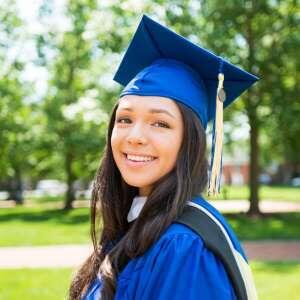 A smiling graduate in a blue cap and gown stands in a green park, capturing a joyful moment of achievement.