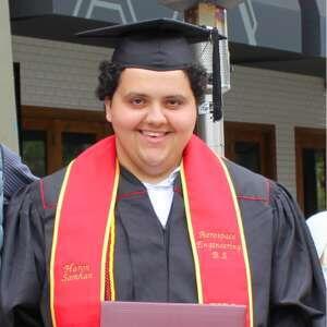 A smiling graduate in cap and gown holds a diploma, wearing red and gold stoles, celebrating academic achievement at a graduation ceremony.