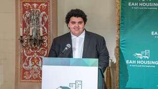 A young man with curly hair stands at a podium, smiling, in front of a backdrop featuring a housing organization.