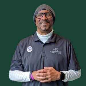 A man in a gray polo shirt and a beanie smiles warmly while standing against a green background, hands clasped in front.