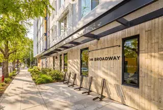 A modern building with a wooden façade and large windows, featuring a sign that reads "ON BROADWAY," set alongside a tree-lined sidewalk.