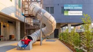 A child in a red jacket enjoys sliding down a large, twisting metallic slide in a modern play area surrounded by greenery.