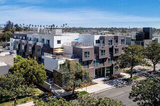 Modern multi-story apartments with balconies, surrounded by trees and sidewalks, under a clear blue sky. Urban living in a sunny setting.