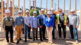 A group of professionals in hard hats stands together at a construction site, smiling and posing in front of a frame structure.