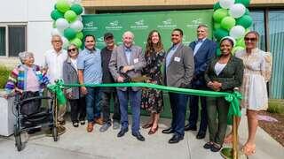 A diverse group of people gathers for a ribbon-cutting ceremony, celebrating a new facility with green balloons and smiles.