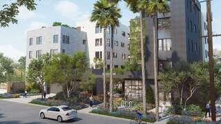 Modern multi-story building surrounded by greenery and palm trees, featuring a welcoming entrance and vibrant landscaping. Pedestrians and cyclists nearby.