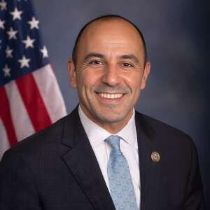 A smiling individual in a formal suit with a light blue tie stands in front of an American flag backdrop, exuding approachability and confidence.