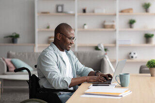 A man in a wheelchair works on a laptop at a desk, surrounded by plants and a cozy living space, exuding focus and productivity.