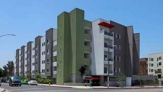 Modern multi-story buildings with a mix of green accents and red highlights, set on a sunny street with parked vehicles.