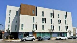 A modern multi-story building with a mix of white and brown facades, featuring large windows and minimal landscaping. Several parked cars are visible.