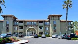 A three-story building with a stucco facade, arched entrance, and palm trees, surrounded by parked cars and vibrant landscaping.