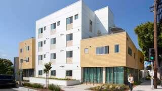 Exterior view of The Laurel, a modern multi-story affordable housing community with white and tan paneling, large windows, and ground-floor retail along a city street.