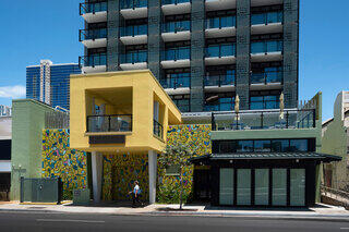 A modern architectural scene featuring a striking yellow building with a mural next to a sleek multi-story structure against a clear blue sky.