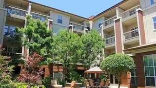 A bright courtyard features lush trees, comfortable seating, and an umbrella-covered lounge area surrounded by multi-story buildings.