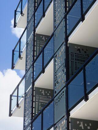 A modern building features a geometric façade with balconies, accented by patterned metal screens and large glass panels against a blue sky.