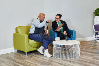 A 62+ male resident and a female real estate management staff member are engaged in a lively conversation on colorful chairs, sharing smiles and ideas over a coffee table in a cozy modern space.