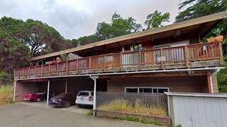 A two-story wooden house features a spacious balcony, surrounded by greenery with parked cars below, reflecting a rustic, peaceful setting.