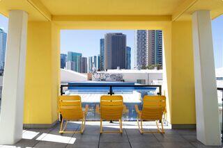 Sunny urban balcony with three yellow lounge chairs overlooking a city skyline and a clear blue pool. Bright and inviting atmosphere.