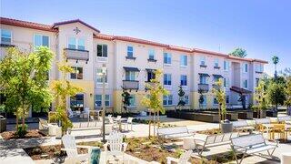 Exterior view of a modern three-story affordable housing community with landscaped courtyard seating and shaded gathering areas.