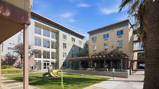A modern residential complex features two buildings with large windows, a shaded patio, and a grassy area alongside a playground.