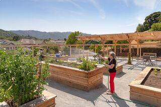An older adult resident waters a rooftop garden with raised beds, surrounded by greenery and mountains, as another person works in the background.