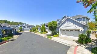 A quiet residential street features charming houses with well-kept lawns under a clear blue sky, creating a peaceful suburban atmosphere.