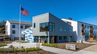 Lightfighter Village features a mid rise affordable housing community by EAH Housing with modern exterior, large windows, and illuminated entry in a residential neighborhood at dusk