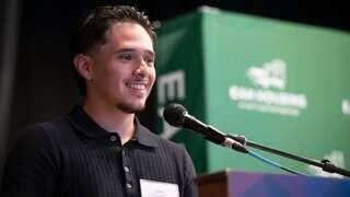 Cesar Gutierrez smiles while speaking at a podium during a scholarship or community event, with an EAH Housing backdrop behind him.