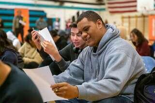 Students sit together in a school gym reviewing papers and notes, engaged in a scholarship or academic event.